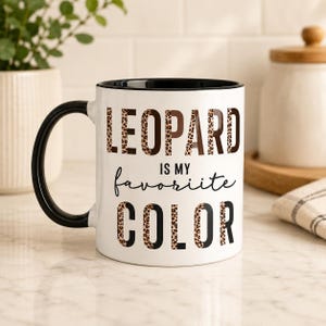 May include: A white ceramic mug with a black handle and interior rim. The front features the text LEOPARD IS MY favoriite COLOR in a mix of bold block letters with a brown leopard print pattern and black script lettering.