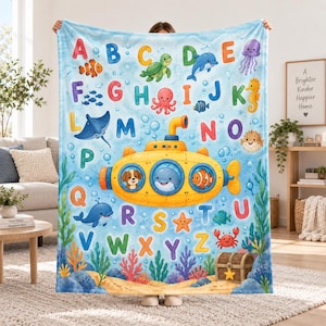 May include: A light blue fleece blanket featuring a colourful alphabet from A to Z, decorated with various sea creatures, a yellow submarine with a puppy, a whale, and a clownfish in the portholes, and a treasure chest on the sandy ocean floor.