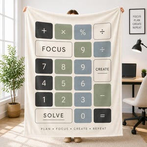 May include: A cream-colored throw blanket featuring a large calculator keypad graphic in shades of charcoal, sage green, and slate blue. The design includes numbers, mathematical symbols, and the words FOCUS, CREATE, and SOLVE, with PLAN • FOCUS • CREATE • REPEAT printed at the bottom.