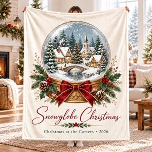 May include: A cream-coloured fleece blanket featuring a large central snow globe illustration with a winter village scene, a stone bridge, and pine trees. The base is decorated with a red bow, pine cones, and holly. Text reads: Snowglobe Christmas, Christmas at the Carters, 2026.