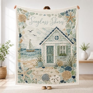May include: A cream-coloured throw blanket featuring a coastal scene with a beach cottage, a sailboat, and seagulls. The border is decorated with various seashells and sea glass in shades of blue, green, and tan. The text Seaglass Shores is printed.