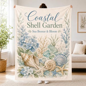 May include: A cream-coloured throw blanket featuring the text Coastal Shell Garden and Sea Breeze & Bloom. The design includes watercolour illustrations of blue hydrangea flowers, various seashells, starfish, coral, and sea glass in soft tones.
