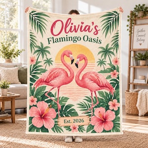 Könnte beinhalten: Eine cremefarbene Fleecedecke mit zwei rosa Flamingos, die sich vor einer gelben Sonne gegenüberstehen. Der Text lautet Olivia's Flamingo Oasis und Est. 2026. Das Design zeigt grüne Palmenblätter und große rosa Hibiskusblüten.