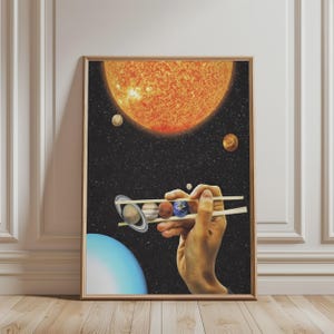 May include: A framed art print featuring a surreal space scene where a hand holds a row of planets between wooden chopsticks. The background is a dark starry sky with a large orange sun at the top and a blue planet at the bottom corner.
