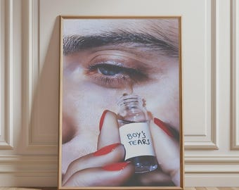 May include: A framed wall art print featuring a close-up of an eye with a tear falling into a small clear glass bottle held by fingers with bright red nail polish. The bottle has a white label with the handwritten text Boy's Tears.