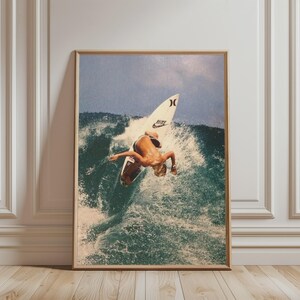 May include: A framed wall art print depicting a surfer riding a wave on a white surfboard with a Nike logo. The surfer is positioned mid-turn on the crest of a breaking ocean wave, with white sea foam spraying around the board in the water.