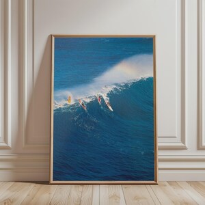 May include: A framed vertical wall art print featuring an aerial view of multiple surfers riding a large, deep blue ocean wave. The crest of the wave shows white sea foam and a faint rainbow mist, with surfers on colorful boards navigating the water.