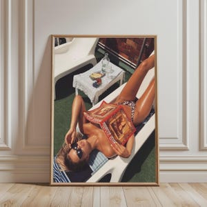 May include: A framed wall art print depicting a person lounging on a white plastic chair outdoors, wearing leopard print swimwear and black sunglasses while reading a red magazine. A small white table with a glass bottle and fruit sits nearby.