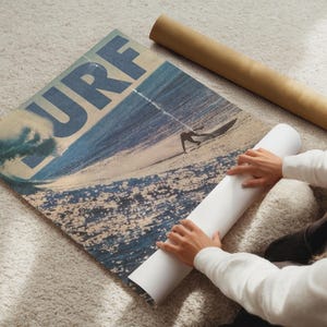 May include: A rectangular poster featuring a large blue wave and a surfer on a sandy beach with the word SURF printed in bold blue letters across the top. The poster is partially rolled up, revealing a white underside and a brown cardboard tube nearby.