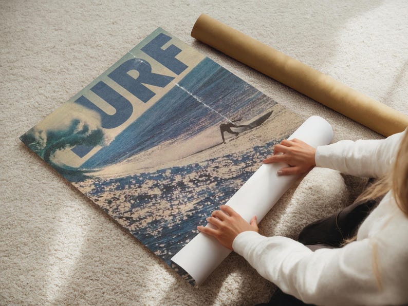 May include: A rectangular poster featuring a large blue wave and a surfer on a sandy beach with the word SURF printed in bold blue letters across the top. The poster is partially rolled up, revealing a white underside and a brown cardboard tube nearby.