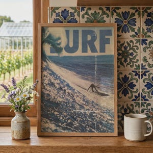 May include: A framed rectangular wall art print featuring a vintage-style graphic of a person surfing a large ocean wave. The word SURF is printed in large blue capital letters at the top. The print is encased in a light-colored wooden frame.