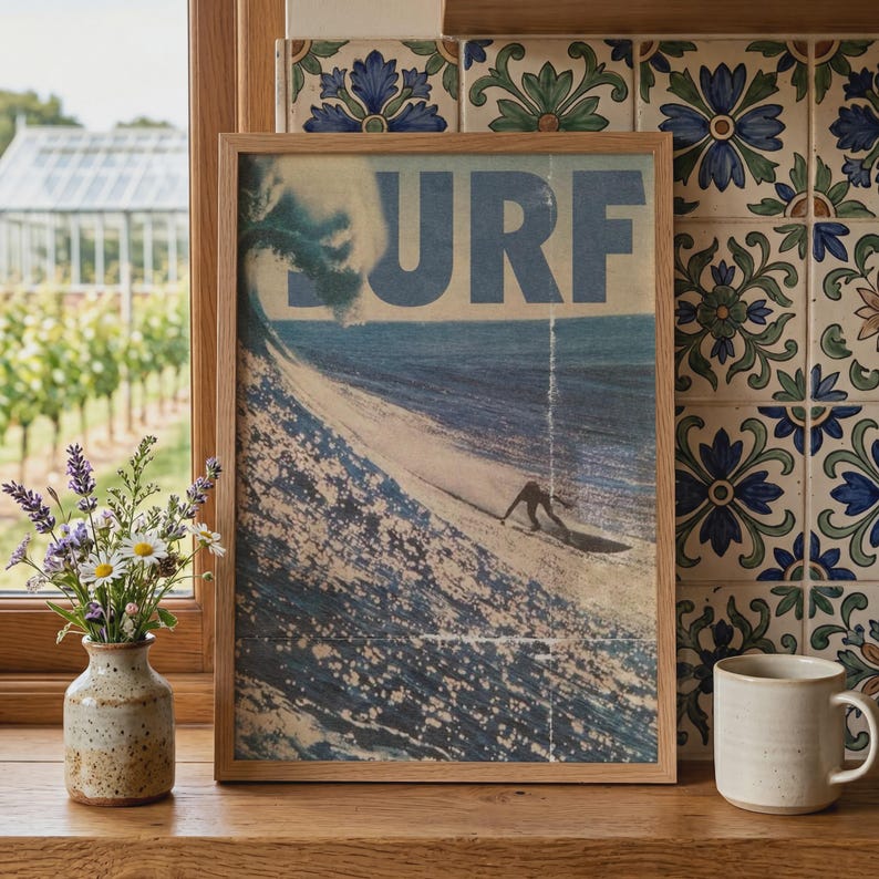 May include: A framed rectangular wall art print featuring a vintage-style graphic of a person surfing a large ocean wave. The word SURF is printed in large blue capital letters at the top. The print is encased in a light-colored wooden frame.