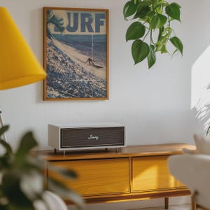 May include: A rectangular white speaker with a dark grey front grille featuring the word Jimmy in white script. The speaker sits on a wooden cabinet below a framed vertical poster depicting a large ocean wave and the word SURF in blue letters.