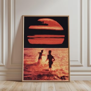 May include: A rectangular wall art print featuring a split composition. The top section displays a large, textured red sun against a solid black background. The bottom section shows two silhouettes of figures running through shallow water.