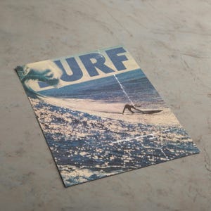 May include: A rectangular print featuring a blue ocean wave and a surfer on a board. The word SURF is printed in large, bold, blue capital letters across the top. The image displays a textured, weathered appearance with a visible vertical crease.
