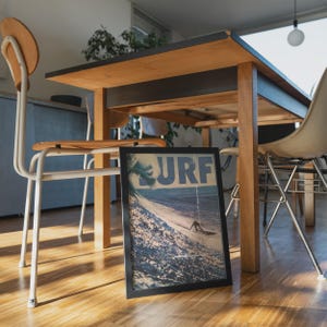 May include: A rectangular framed print featuring a blue and white graphic of a large ocean wave and a surfer with the text SURF in bold blue letters at the top. The framed artwork is positioned on a wooden floor beneath a wooden dining table.
