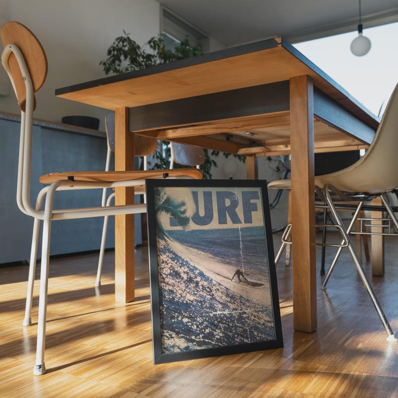 May include: A rectangular framed print featuring a blue and white graphic of a large ocean wave and a surfer with the text SURF in bold blue letters at the top. The framed artwork is positioned on a wooden floor beneath a wooden dining table.