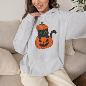 Könnte beinhalten: Ein hellgrauer Kapuzenpullover mit einem Grafikdruck einer schwarzen Katze mit grünen Augen, die in einem geschnitzten orangefarbenen Kürbis sitzt. Der Hoodie verfügt über eine Kängurutasche vorne und weiße Kordelzüge am Halsausschnitt.