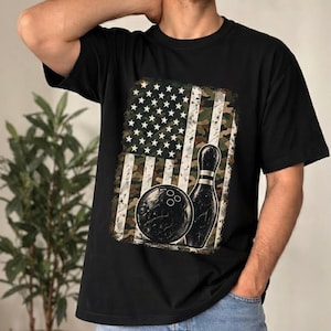 May include: A black t-shirt featuring a rectangular graphic print. The design displays a stylised American flag with a camouflage pattern overlay, incorporating a black bowling ball and a single white bowling pin positioned in the foreground.