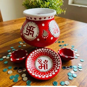 Op de afbeelding: Een rood beschilderde aarden pot en bijpassend bordje, versierd met witte stippen, kleine ronde spiegeltjes en de symbolen Swastika en Maa in rood en wit. Twee kleine rode aarden olielampjes staan ernaast, omringd door losse ronde spiegeltjes.