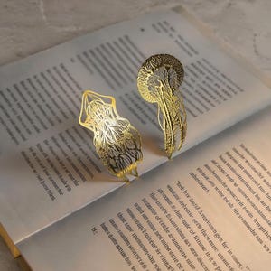 Può includere: Due accessori per capelli in metallo dorato con intricati disegni in filigrana tagliati al laser. Un pezzo presenta una forma simmetrica allungata, mentre l'altro mostra una struttura arrotondata a ventaglio con delicati elementi pendenti.