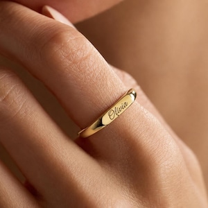 May include: A polished gold band ring featuring a smooth, rounded surface with the name Olivia engraved in a cursive script font. The ring has a reflective metallic finish and a simple, clean design that wraps around the finger.