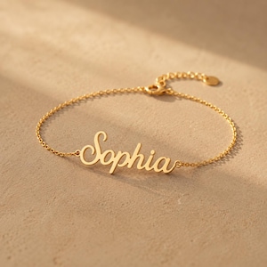 May include: A gold-toned metal bracelet featuring a delicate chain and a central nameplate with the name Sophia written in a cursive script font. The bracelet includes a small circular clasp mechanism at the end of the chain for secure fastening.