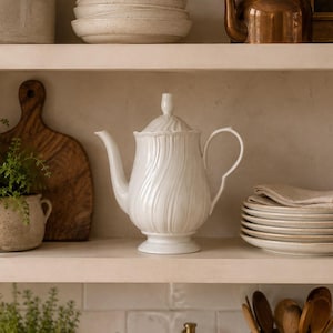 May include: A white ceramic teapot with a vertical swirl pattern, a pedestal base, and a matching lid. The teapot features a curved spout and a handle, positioned centrally on a shelf among other kitchen items like bowls and a copper kettle.