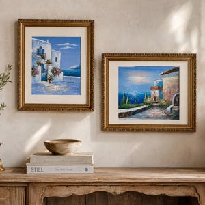 May include: Two framed oil paintings displayed on a wall. The left painting depicts a white Mediterranean building with blue accents and flowers. The right painting shows a coastal landscape with stone structures, greenery, and a blue sea view.