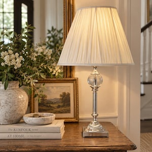 May include: A table lamp featuring a square silver base, a slender metallic stem, and a clear glass sphere accent. The lamp is topped with a white, pleated, conical fabric lampshade that emits a warm, soft glow when illuminated.