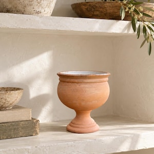 Puede incluir: Un cuenco con pedestal de color terracota con cuerpo redondeado, borde acampanado y base circular ancha. El interior del cuenco presenta un acabado blanco liso, mientras que el exterior muestra una textura mate y terrosa con sutiles variaciones.