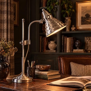 May include: A brushed metal desk lamp with a tiered, conical shade and an adjustable arm. The lamp features a circular base, a vertical support rod, and a light-colored power cord. The shade has a series of small circular ventilation holes.