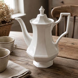May include: A white ceramic teapot with a faceted, octagonal body, a pointed lid, and a geometric handle. The teapot features a beaded detail around the midsection and stands on a pedestal base, showcasing a glossy finish and a curved spout.