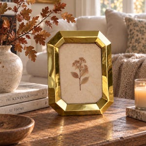 May include: A polished gold-toned metal picture frame with a faceted, octagonal border. The frame holds a light-colored, textured matting featuring a single pressed botanical specimen with small brown flowers and leaves centered within the glass.
