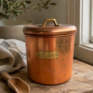 May include: A cylindrical copper canister with a matching lid, featuring a brass handle and a brass plaque engraved with the word SUGAR. The canister has a polished metallic finish and is accompanied by a small, light-colored wooden scoop.
