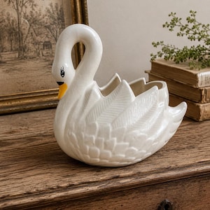 Puede incluir: Una maceta de cerámica blanca con forma de cisne, acabado brillante, cuello largo y curvado, y texturas de plumas moldeadas en el cuerpo. El cisne presenta un pico pintado de amarillo y detalles oculares en negro con una cavidad abierta.