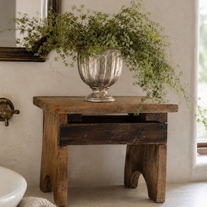 May include: A small, rectangular wooden stool with a dark, weathered finish and four sturdy legs. Resting on top is a metallic, silver-toned pedestal vase with a fluted design, filled with a lush, trailing green plant with delicate foliage.