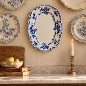 May include: An oval ceramic serving platter featuring a white center and a blue floral pattern along the scalloped rim. The platter is displayed on a wall alongside other decorative plates, with a candle in a brass holder nearby.