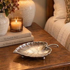 May include: A silver-toned metal dish shaped like a leaf with a stem handle and three small ball feet. The surface features embossed vein details and a scalloped edge, providing a shallow basin for holding small items on a flat surface.