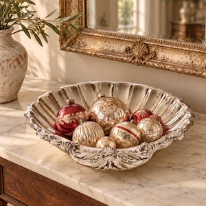 May include: A silver-toned scalloped bowl with ornate embossed detailing along the rim, filled with a collection of round glass ornaments in shades of red, gold, and mercury glass with various textures and painted leaf patterns.