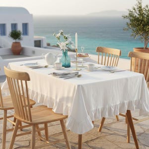 May include: A white rectangular tablecloth with a ruffled edge draped over a table. The table is set with a white teapot, white cups and saucers, a tall thin candle in a gold holder, and a glass vase containing white flowers on a patio.