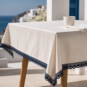 Pode incluir: Uma toalha de mesa quadrada em tom branco sujo com uma borda de renda de crochê azul-marinho escuro. A toalha está sobre uma mesa de madeira, com uma caneca de cerâmica a condizer e um guardanapo branco dobrado sobre a superfície.