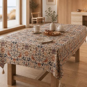 Peut inclure: Une nappe rectangulaire sur fond crème ornée d'un motif répétitif d'oiseaux orange, de fleurs bleues et orange, et de feuillage vert. Les bords du tissu sont agrémentés de petites franges de couleur claire à chaque coin.