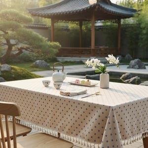 May include: A beige tablecloth with a repeating pattern of small blue circles and a white tassel fringe trim. On the table sits a white teapot, two small white bowls, a rectangular tray with three small cakes, and a white vase with white orchids.