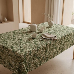 Può includere: Una tovaglia rettangolare caratterizzata da un fitto motivo ripetuto di piccoli fiori bianchi su sfondo verde scuro. La tavola è apparecchiata con una teiera in ceramica bianca, due piccole ciotole bianche, un piatto bianco e un tovagliolo di lino beige piegato.