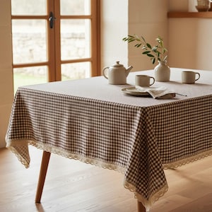 Può includere: Una tovaglia rettangolare con motivo a quadretti vichy marroni e bianchi, rifinita con un delicato bordo in pizzo color crema. La tovaglia copre un tavolo apparecchiato con una teiera in ceramica, due tazze e un piccolo vaso.