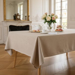 May include: A rectangular beige tablecloth with a visible woven texture draped over a table. The table is set with a white teapot, a white teacup, a glass vase containing white and pink roses, and a tiered stand holding small colorful pastries.