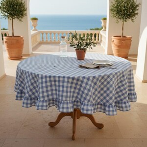 Peut inclure: Une table ronde en bois recouverte d'une nappe à carreaux bleus et blancs avec un bord à volants. Sur la table se trouvent une bouteille en verre transparent, une petite plante verte en pot et une assiette blanche avec une serviette pliée.
