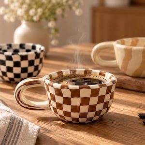 Può includere: Tre tazze in ceramica con manici arrotondati. Una tazza presenta un motivo a scacchi bianco e nero, un'altra un motivo a scacchi marrone e bianco riempito di caffè fumante, e la terza un motivo a strisce ondulate beige e bianco.