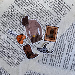 Op de afbeelding: Een verzameling van vijf gestanste stickers met een getatoeëerde ontblote torso met een cowboyhoed, een bruine cowboylaars met vlamdetails, een schedel van een longhorn, een postzegel met een laars en een hartvormige lolly met de tekst Sucker for Cowboys.